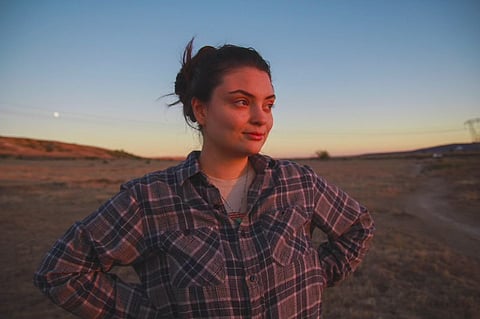 A photo of Ayşenur Ezgi Eygi standing outdoors with her hands on her hips, looking to the side of the camera, her face lit by the setting sun. Her hair is pulled up and she's wearing a flannel shirt.