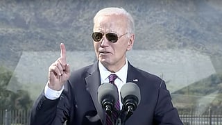 U.S. President Joe Biden stands outdoors, wearing a dark suit, tie, and sunglasses, with his right hand raised and index finger pointing upward as he speaks. Two microphones are positioned in front of him, and a blurred mountain landscape is visible in the background.