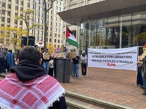 A group of people gather in a plaza for a pro-Palestine rally. A person at a podium speaks, while others hold signs with messages supporting Palestinian liberation. A large banner behind them reads, "Struggle for Liberation! Defend People's Struggle! ILPS." A Palestinian flag is displayed prominently.
