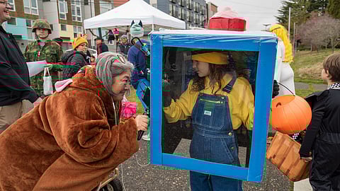 A child dressed in overalls and a yellow hat stands inside a homemade blue costume resembling a vending machine, complete with a clear window displaying items. An older woman in a brown animal-themed costume interacts with the child through the "vending machine" window.