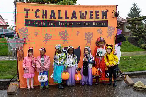 A group of children in Halloween costumes stand in front of an orange backdrop labeled "T'Challaween: A South End Tribute to Our Heroes," decorated with Black Panther symbols and Halloween icons. The kids wear various costumes, including a unicorn, princesses, an astronaut, and a Star Wars pilot, holding trick-or-treat bags and pumpkin buckets.