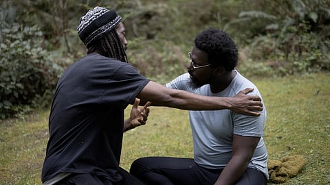 Two men are sitting on grass in a peaceful, wooded area, engaged in a serious conversation. One man, wearing a beanie, gestures with open hands while holding the other’s shoulder, who looks back attentively.