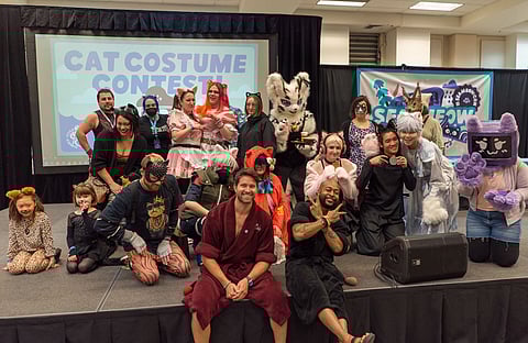 A large group of participants posing on stage for a "Cat Costume Contest" at a convention. People of all ages are dressed in various cat-inspired costumes, including onesies, furry suits, cat ears, and face paint. Some are in elaborate outfits with props, while others wear simpler cat accessories. Two hosts in robes sit at the front, smiling and posing. A large event logo is displayed on the screen behind them.