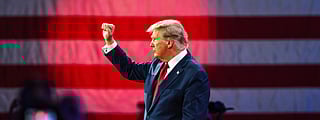 Donald Trump in a suit and red tie stands on a stage with his fist raised, in front of a large American flag backdrop. He faces sideways toward an audience, who are partially visible, with some taking photos.