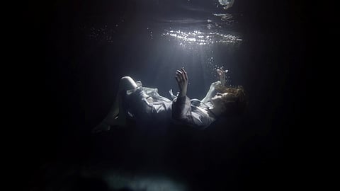 A person in a light-colored outfit floats in dark water, illuminated by a soft beam of light from above. Their body appears weightless, limbs gently raised, with small air bubbles rising. The overall scene is quiet and ethereal, with contrasting light and shadows creating a sense of calm and mystery.