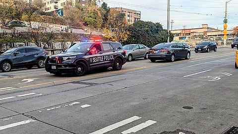 Seattle Police SUV with flashing lights is stopped in the middle lane of a multi-lane street with other vehicles lined up behind it. Surrounding area includes trees, buildings, and parked cars along the roadside. The road shows visible cracks and worn lane markings.