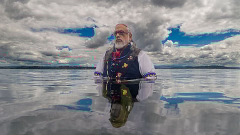 An older man with long gray hair and a beard stands in calm water up to his chest, gazing thoughtfully into the distance. He wears traditional Indigenous attire with colorful beadwork, including a vest, necklaces, and armbands, under a cloudy sky with patches of blue. His reflection is visible in the water.