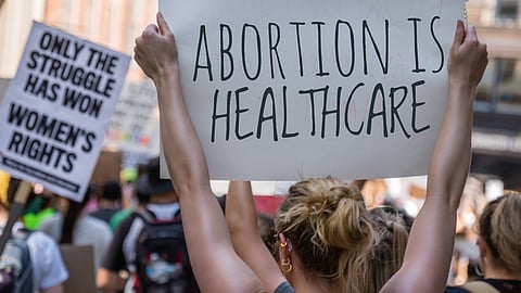 A person holding a large sign that reads "ABORTION IS HEALTHCARE" at a protest. In the background, other demonstrators carry signs, including one that says "ONLY THE STRUGGLE HAS WON WOMEN'S RIGHTS." The scene is crowded, and participants wear casual clothing.