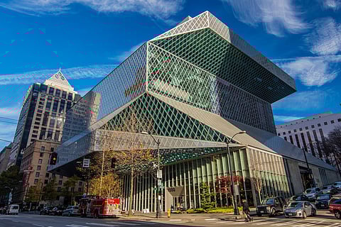 Street view of the Seattle Central Library, a modern building with an angular, geometric design featuring large glass windows in a diamond-patterned grid. The structure has several stacked and offset sections.