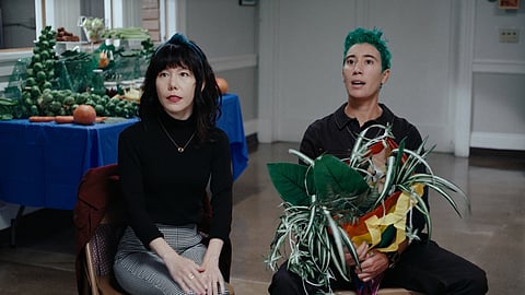 A film still shows Yvette on the left, sitting with her hands on her knees and looking at something beyond the camera, and Syd, right, also looking at something beyond the camera and holding a large plant.