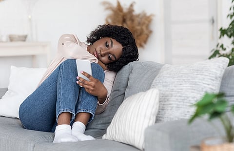 A black woman who looks sad is curled up on the couch, her arms around her legs, looking at her phone.