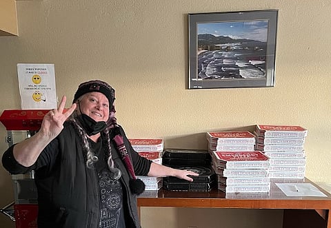 A smiling woman wearing a winter hat and scarf flashes a peace sign while standing next to a stack of pizza boxes on a wooden counter. A landscape photo hangs on the wall above the pizzas, and a sign with a smiley face is partially visible on the left.