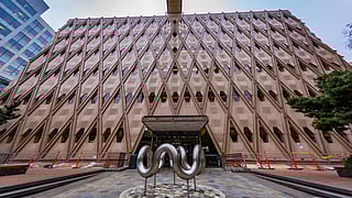 The King County Administration Building, featuring a unique geometric facade with diamond-shaped patterns. In front of the entrance stands a modern metal sculpture with curved shapes, surrounded by a tiled plaza and bordered by trees. Bright orange safety barriers are visible nearby.
