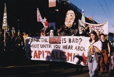 Protesters march behind a banner reading "You think the WTO is bad? Wait until you hear about CAPITALISM?" adorned with a black and red star. Behind them are puppets and other banners.