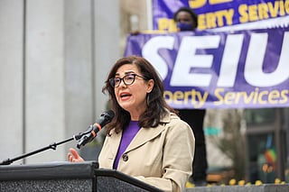 A woman speaks at a podium during a rally, wearing glasses, a beige coat, and a purple shirt, with a microphone in front of her. Behind her, a banner reads "SEIU Property Services" in bold white and yellow text.