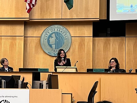 A speaker stands at a podium in a formal meeting room with the "City of Seattle" seal prominently displayed behind them. Two women are seated on either side, one on the left looking toward the speaker and the other on the right smiling