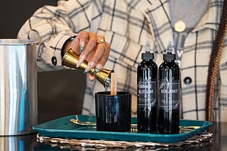 Colina Bruce pouring ingredients for candle-making. Next to her hand sits two bottles, one reads, "Orange Blossom" and the other "Bergamot."