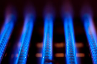 Close-up of blue flames burning in a gas furnace, showing multiple parallel gas burners glowing brightly in a dark environment. The vibrant blue color contrasts with the dark background.