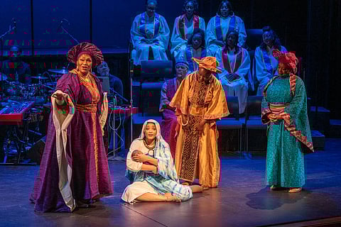 Four performers on stage in colorful, traditional African-inspired costumes during a theater performance. A woman in a blue robe is kneeling, holding a swaddled baby, surrounded by three standing women in rich purple, gold, and turquoise outfits. A choir in white robes is seated in the background under blue stage lighting.