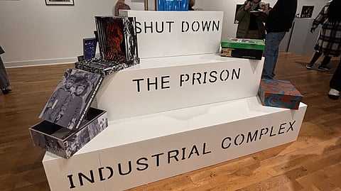 An art exhibit installation featuring a tiered white platform with bold black text reading "SHUT DOWN THE PRISON INDUSTRIAL COMPLEX." Various artistic pieces, including paintings and boxes with creative designs, are displayed on the platform. Visitors are seen engaging with the exhibit in a gallery space with framed artworks on the walls.