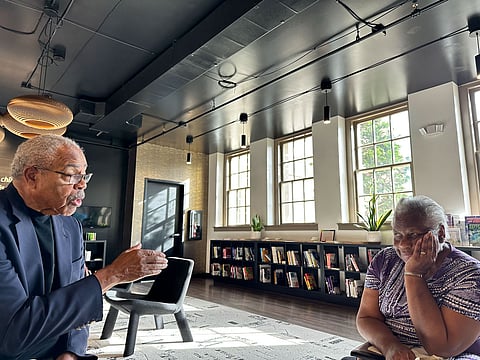 An elderly Black man with glasses, wearing a dark suit, is speaking animatedly with his hand gesturing, while an elderly Black woman in a purple patterned shirt listens with her hand resting on her cheek. They are in a modern, well-lit room with large windows, bookshelves filled with books, and plants.
