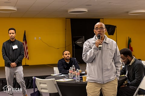 A man speaks into a microphone while standing in a community event setting with a yellow wall behind him. Seated attendees and a presenter in a black jacket are visible, along with materials and water bottles on tables. An American flag and Ethiopian flag are seen in the background.