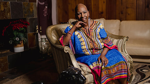 An older Black woman with a shaved head sits on an ornate beige chair in a cozy living room, wearing a vibrant blue and multicolored traditional patterned dress. She smiles warmly, resting her chin on her hand. The background features wood-paneled walls, a stone fireplace, and a poinsettia plant.