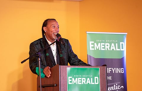 A Black man with long braids and a green patterned jacket speaks enthusiastically at a podium with a microphone. The podium and a banner beside it display the "South Seattle Emerald" logo. The background is a bright yellow wall.