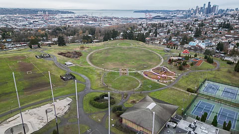 Aerial view of a park with sports fields, playgrounds, and tennis courts, with the city skyline and harbor in the background on an overcast day.