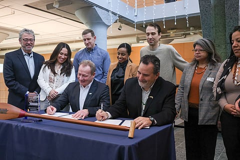 A group of individuals gathers around a table as two men, seated in suits, sign documents during a formal ceremony. A ceremonial paddle and a plaque with the Port of Seattle logo are displayed on the table.