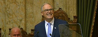 Washington State Governor Jay Inslee delivering the "State of the State Address" at the Washington State House of Representatives on January 14, 2025. He is standing at a podium with a smile, wearing a suit and tie, while another person applauds in the background. A caption at the bottom of the image provides details about the event.