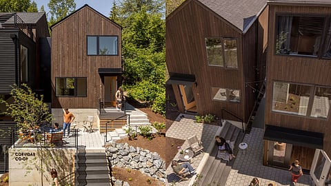 A modern co-housing community with dark wood-clad homes and shared outdoor spaces. Residents interact on patios, stairs, and walkways, surrounded by greenery and landscaping.