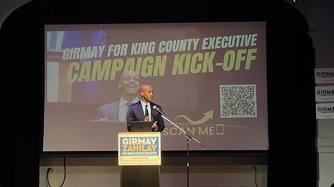 A Black man stands on a stage and addresses a crowd with an image behind him that reads "Girmay for King County Executive Campaign Kick-Off."