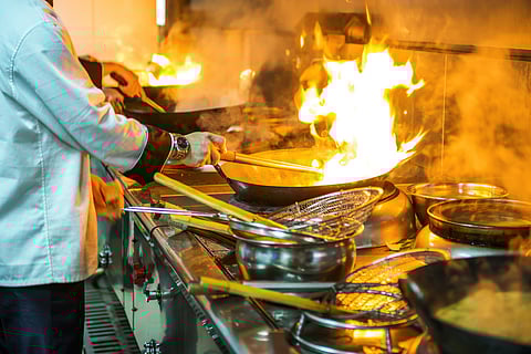 A chef in a white jacket cooks in a professional kitchen, flambéing a dish with tall flames rising from a wok. Various kitchen utensils and pots are visible on the counter.