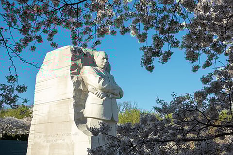 The Martin Luther King Jr. Memorial in Washington, D.C., featuring a towering stone sculpture of Dr. King with arms crossed, framed by blooming cherry blossoms against a clear blue sky. The inscription on the stone reads, "Out of the mountain of despair, a stone of hope."