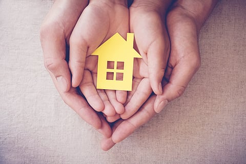 Close-up of two pairs of hands, an adult's and a child's, cradling a yellow paper cutout of a house with windows and a chimney.