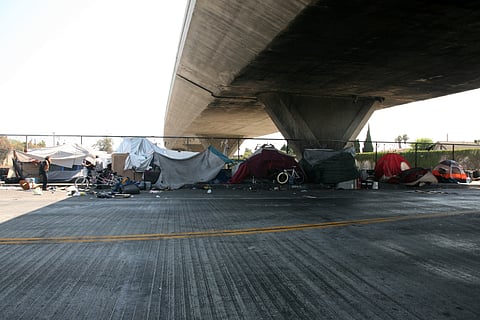 A homeless encampment set up along a fence under a highway overpass.
