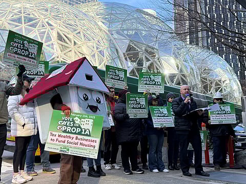 A group of protesters gather outside a modern glass-domed structure, holding signs in support of "Prop 1A" for social housing. One protester is dressed in a house costume with a smiling face. A speaker addresses the crowd with a microphone, while others hold signs reading "Vote Yes on Prop 1A" and "Tax Wealthy Businesses to Fund Social Housing."
