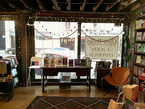 A cozy independent bookstore with wooden floors, exposed wooden ceiling beams, and large front windows adorned with string lights and colorful garlands. A banner in the window reads "Open Books: A Poem Emporium" in reverse.