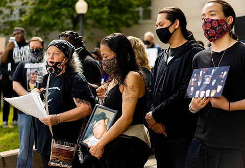 A group of people are standing in a row. One is holding a microphone. One is holding a photo of a young man.