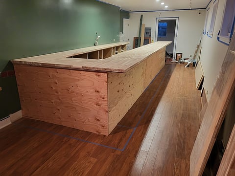 A partially constructed wooden bar with an L-shaped countertop is in the center of a room under renovation.