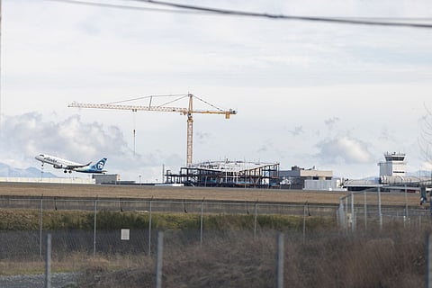 An Alaska Airlines plane is taking off from an airport runway, ascending into the sky. In the background, a large construction crane towers over a partially built structure.