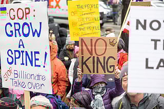 A crowd of protesters gathers on a city street holding various handmade signs. A central sign, held by a person in a purple coat, patterned scarf, and bucket hat, reads "NO KING" in bold red and black letters with a small crown drawn above the word "NO."