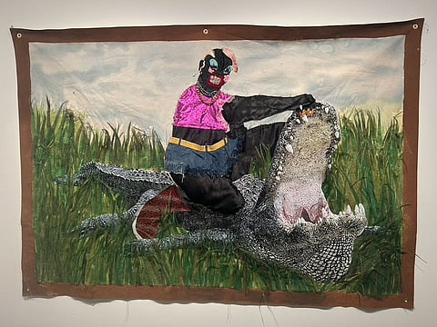 A textile artwork depicting a surreal scene of a figure sitting atop a large alligator in a grassy environment. The figure is dressed in a bright pink top, dark pants, and a golden belt, with a masked face adorned with embellishments. The alligator has its mouth wide open, showcasing sharp teeth and a textured snout.