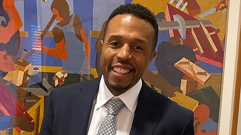 A Black man dressed in a suit stands in front of a Jacob Lawrence artpiece.