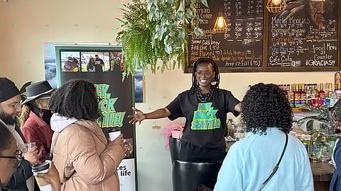 A group of Black individuals gathers inside a café, listening to a speaker standing behind the counter. The speaker wears a "Hey Black Seattle" shirt and gestures while engaging with the audience, who hold cups of coffee.