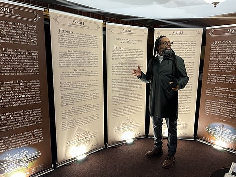 A Black man with long dreadlocks, glasses, and a beard stands in front of tall informational banners about the history of Black churches, gesturing as if speaking.