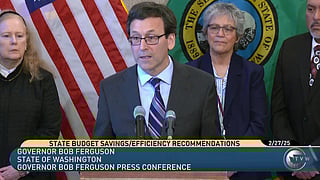 Governor Bob Ferguson speaks at a podium during a press conference on state budget savings, with three individuals standing behind him in front of the U.S. and Washington State flags. A news banner displays the event details, including the date "2/27/25" and the TVW logo.