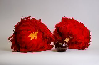 A pair of bright red ʻuliʻuli, traditional Hawaiian feathered gourd rattles, rest on a plain white surface. One rattle has a yellow feather pattern at its center, while the other shows its polished brown wooden handle with a round, glossy end.