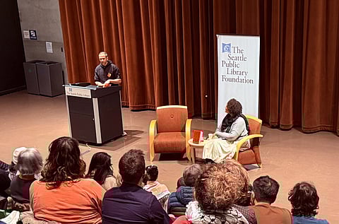 Omar El Akkad (left, at podium) joined Ijeoma Oluo (right, seated) at the Seattle Central Library in conversation about El Akkad's book "One Day, Everyone Will Have Always Been Against This." Audience members are visible in the foreground, listening to El Akkad speak. A large curtain is the backdrop of the conversation.